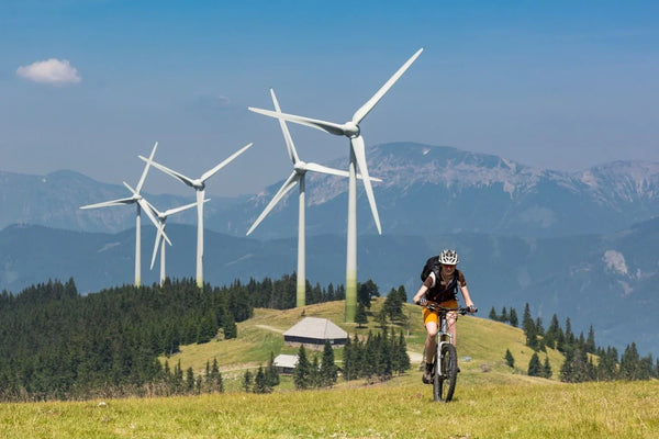 Cycliste casqué à VTT sur un chemin herbeux, devant des éoliennes et un paysage montagneux.
